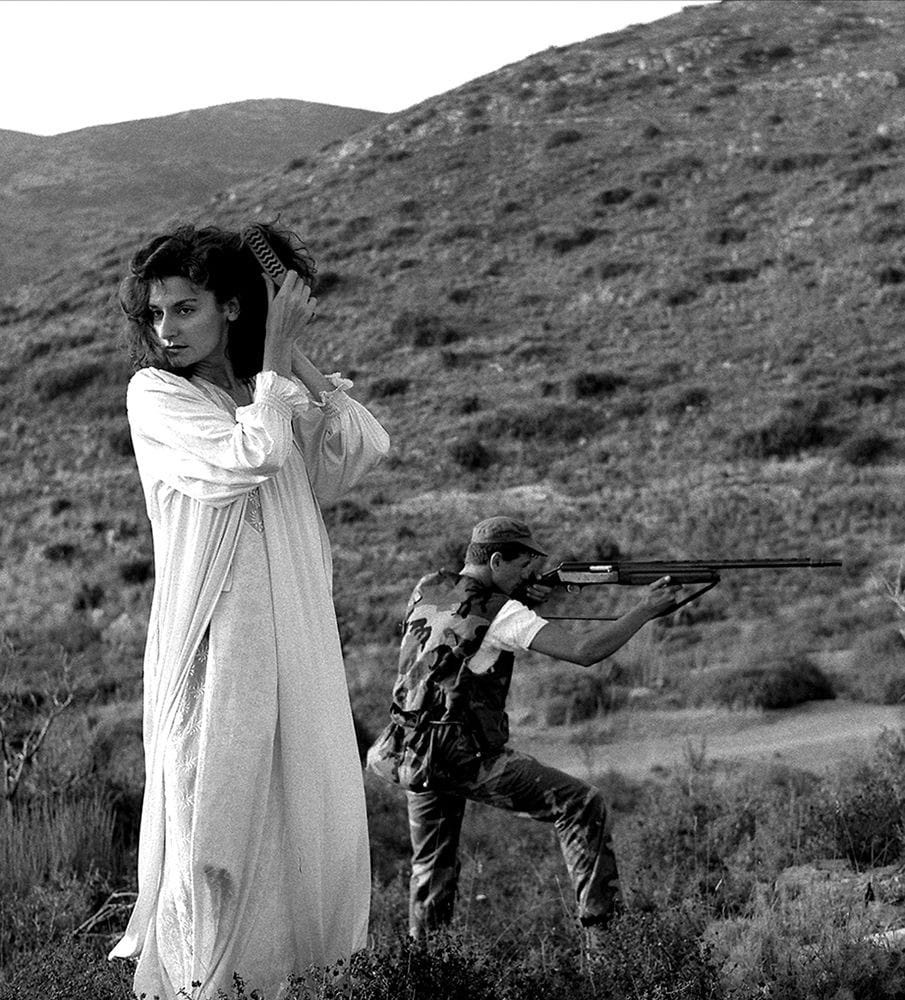 Portrait of woman in nature seductively brushing her hair with hunter in background aiming his rifle.